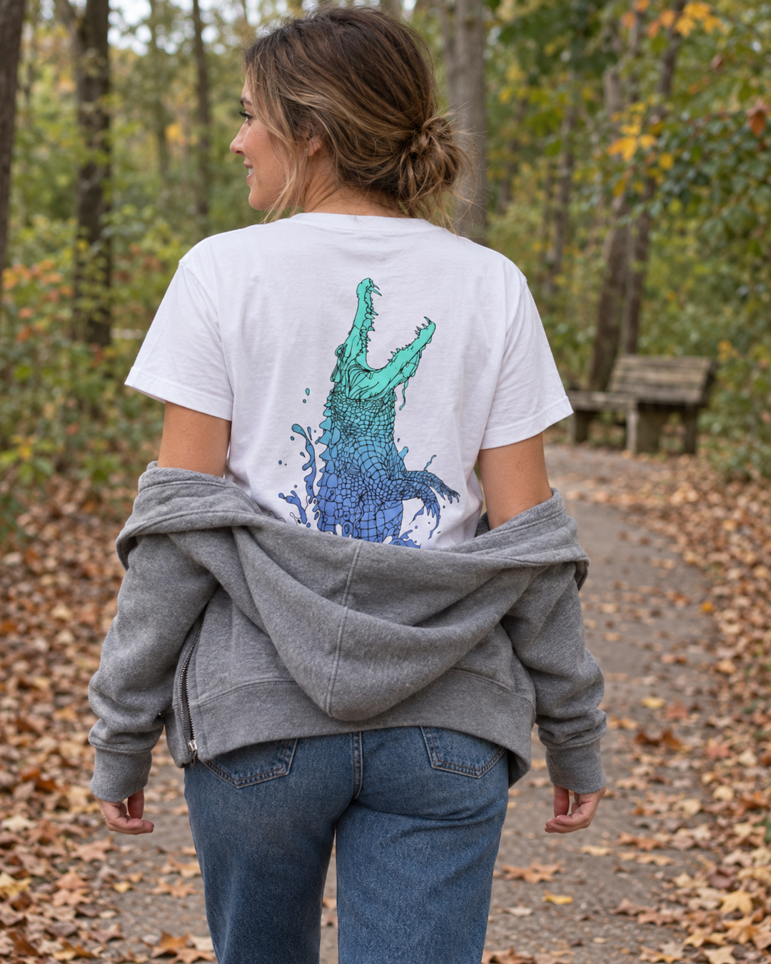 Woman wearing a white Nebula Crocodile graphic tee on an autumn trail, with a grey hoodie slipped below her shoulders to reveal the back print.