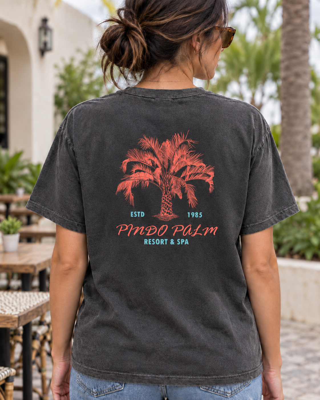 Woman wearing a Pepper Pindo Palm t-shirt outside a cafe, showing the smaller centered back graphic.