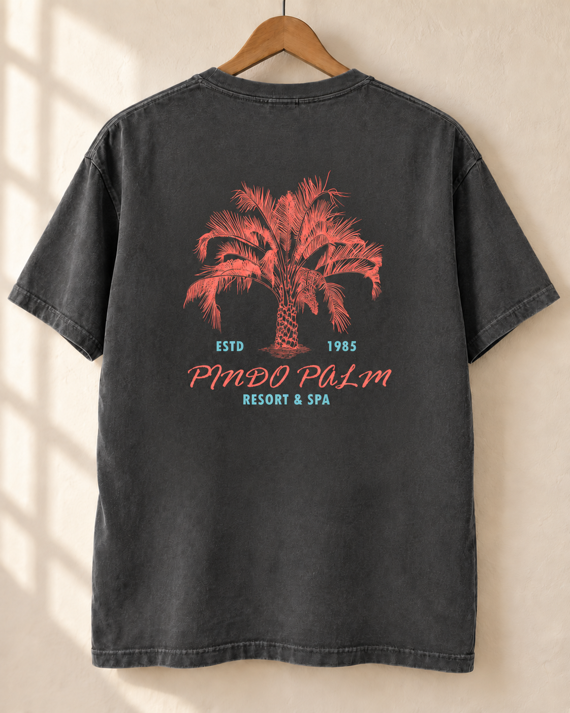 Pepper Pindo Palm t-shirt on a wooden hanger, showing the centered coral palm and turquoise resort lettering on the back.