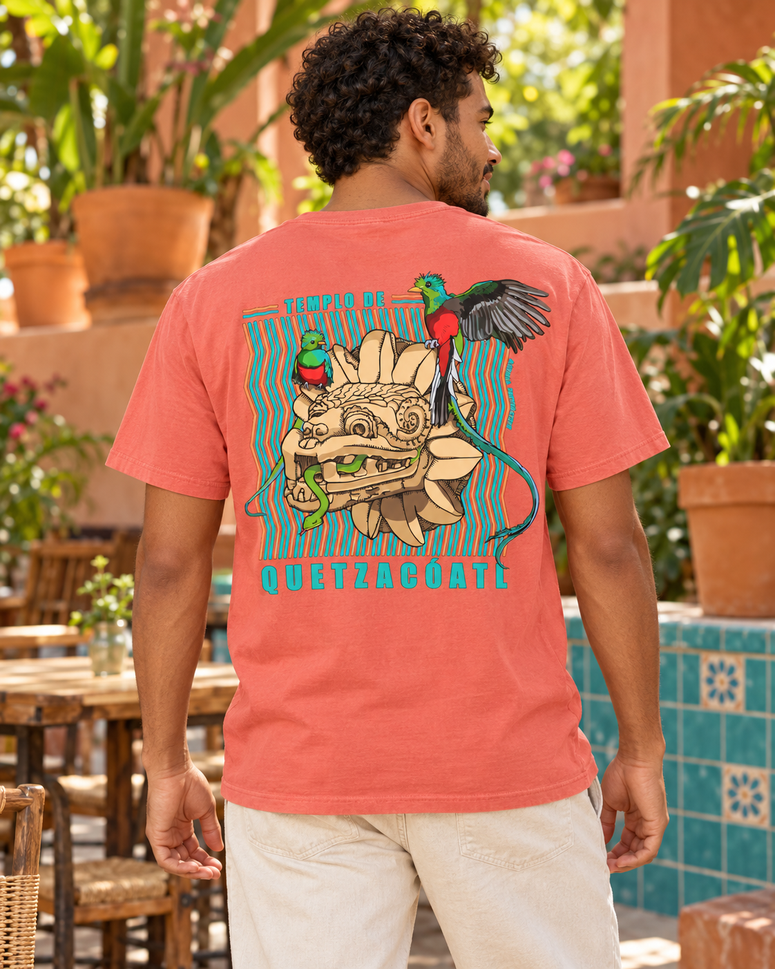 Man wearing the Bright Salmon unisex Quetzalcoatl graphic tee, showing the back print beside turquoise courtyard tiles.