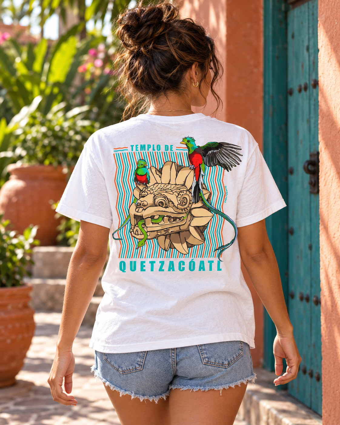 Woman wearing a white unisex Quetzalcoatl graphic tee, showing the colorful back artwork in a tropical courtyard.